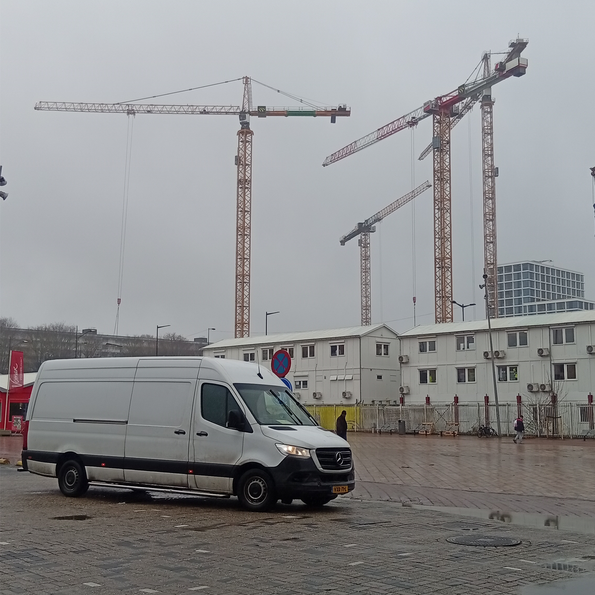 Work in progress  Urban construction site in Amsterdam Zuidoost with cranes, temporary buildings and a van under a grey rainy sky.